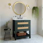 A green single sink bathroom vanity with cane drawer fronts, black and gold accents, and a Quartz top. The vanity is assembled and ready for installation, displayed in a room setting with a round mirror above the sink and a plant in the background.