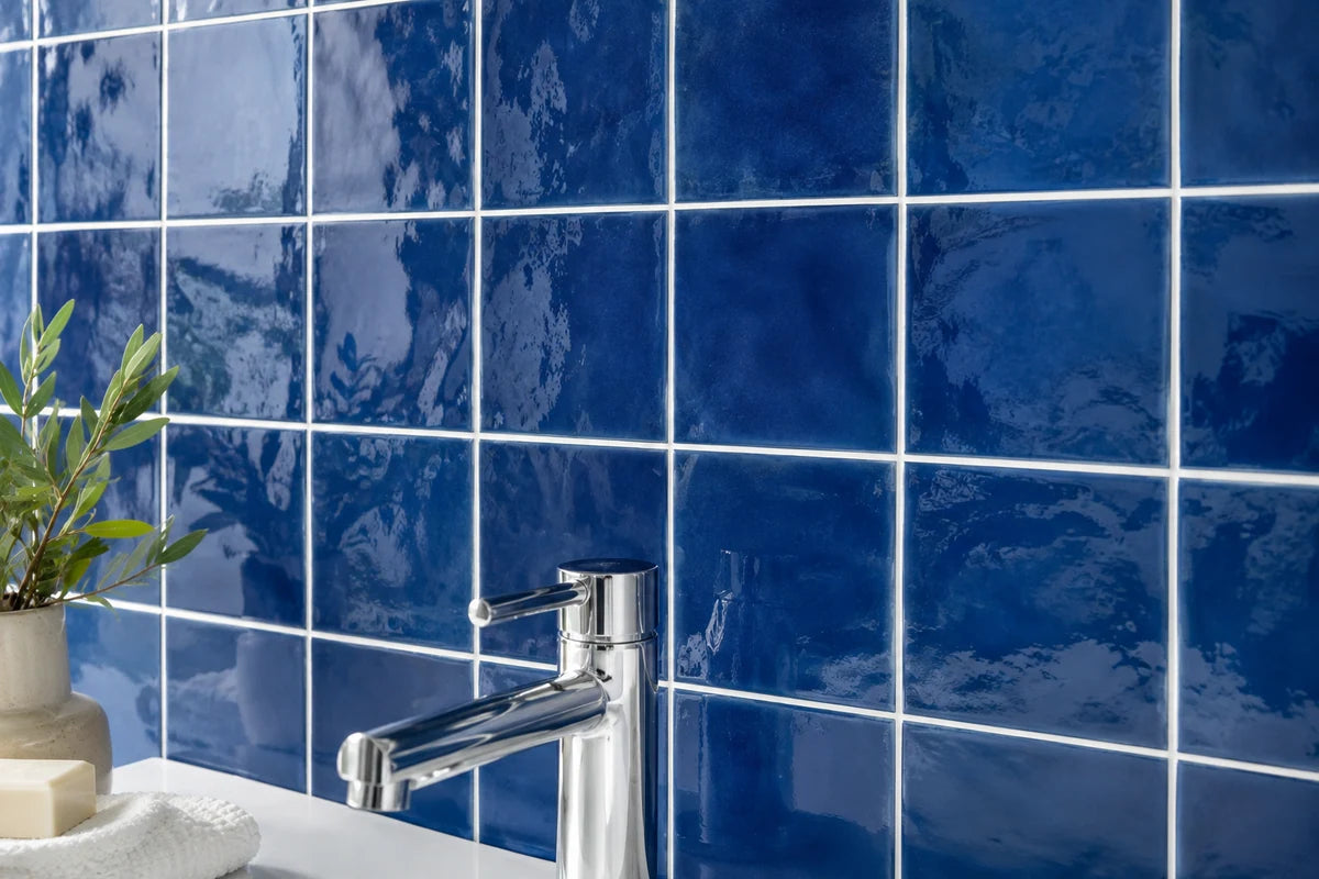 Close-up of cobalt blue glossy ceramic square tiles on a bathroom wall, gently rippled glaze, neat white grout, partial chrome faucet at lower edge